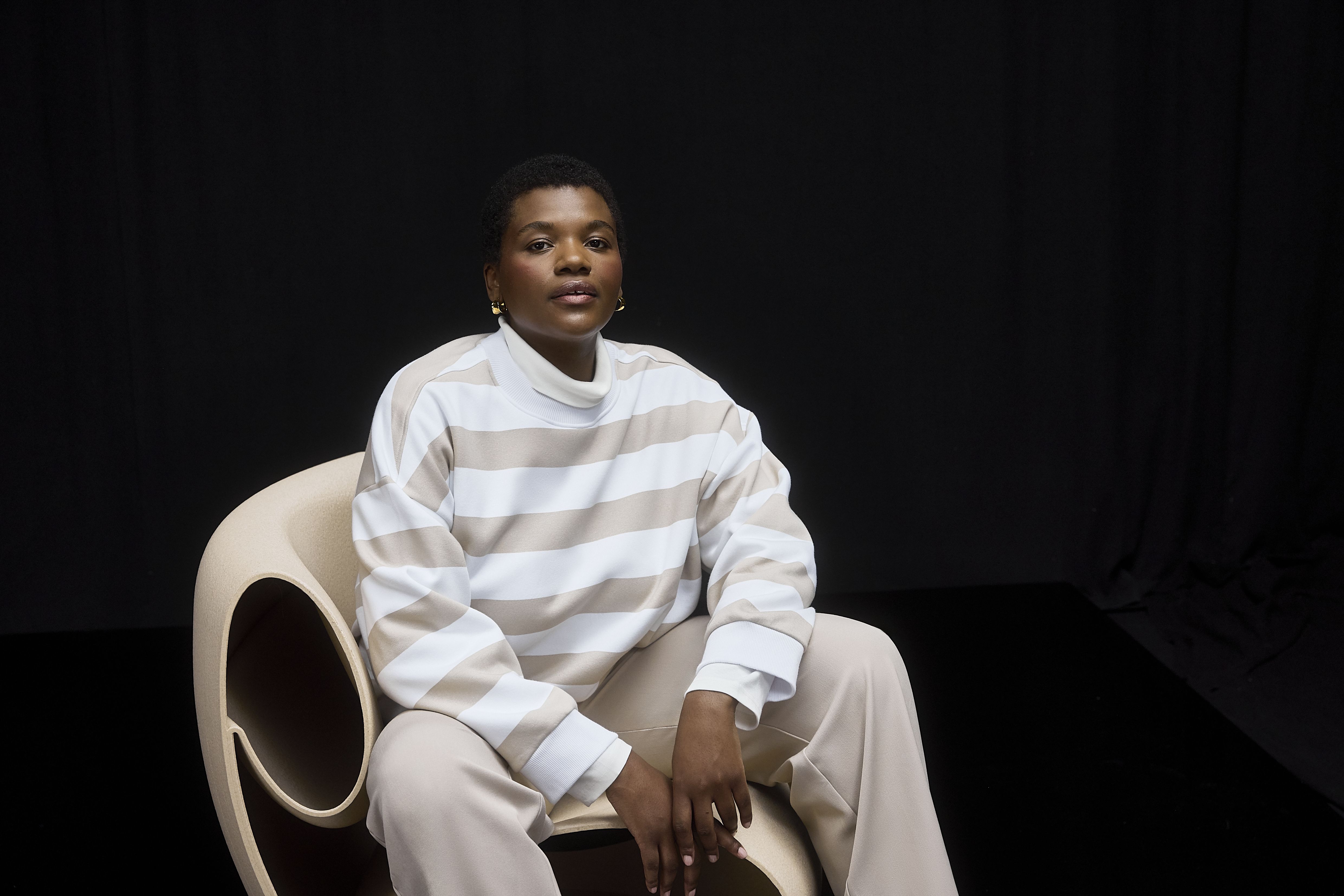 Femme assise, portant un sweat rayé beige et blanc, un pantalon beige et des baskets blanches